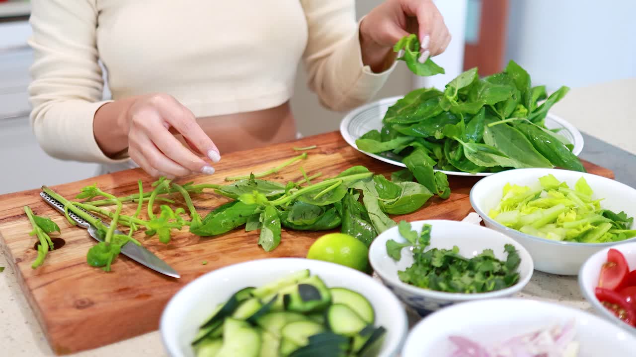 Hands skillfully prepare fresh basil on a wooden board, surrounded by vibrant vegetables in a well-lit kitchen