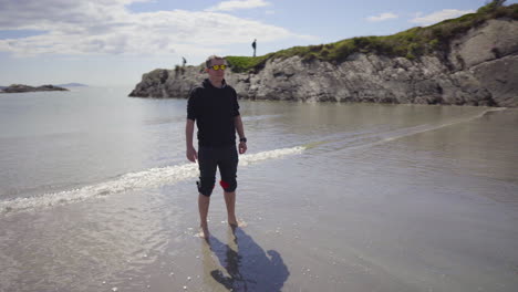 Tiro-En-Círculos-Alrededor-De-Una-Persona-De-Pie-En-La-Playa-De-Arena-Con-Gafas-De-Sol-Con-Olas-Del-Océano-Salpicando-En-4k