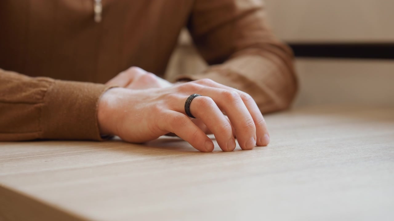 Primo piano delle mani di un uomo appoggiate su una superficie di legno chiaro, le dita si muovono leggermente con un anello nero visibile. Le mani rimangono a fuoco mentre lo sfondo sfuma delicatamente, enfatizzando la texture naturale della pelle.