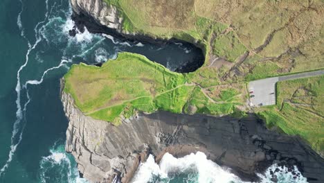 drone shot of the kilkee cliffs of ireland, where the ocean meets the countryside fields and waves are crashing against the tall rugged coastline