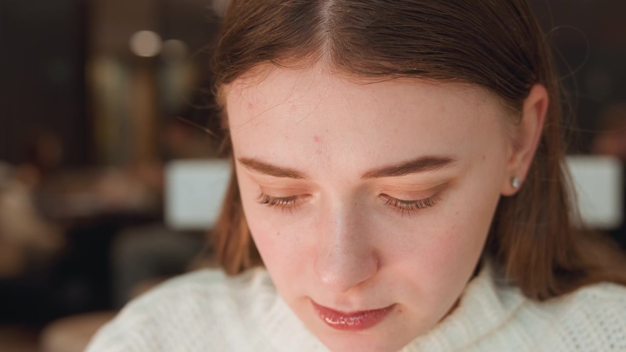 Tight close-up of young woman looking downward with gentle, contemplative expression and subtle gold eyeshadow on upper eyelids; soft warm indoor light casts gentle highlights on freckles