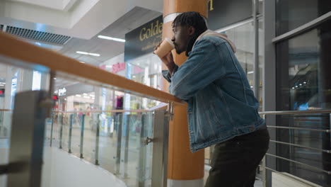 young man in denim jacket and hoodie sipping coffee while leaning on railing in brightly lit shopping mall, surrounded by modern glass architecture, visible storefronts, and relaxed atmosphere of retail space
