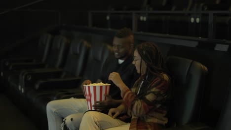 African-American-young-couple-came-to-the-movie-theatre-watching-film,-taking-popcorn-from-bucket