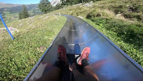 pov shot of a man holding onto a small toboggan, sliding down a long metal slide through the beautiful scenery of the swiss alps