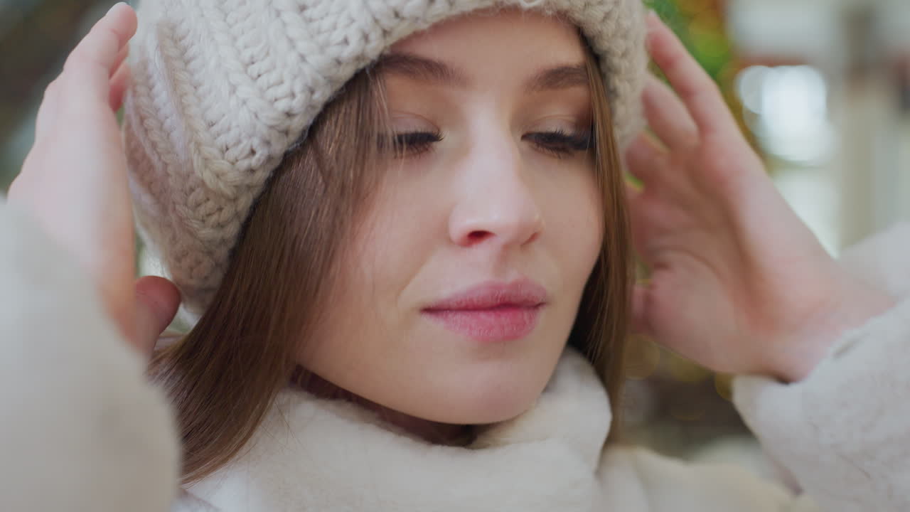 Close-up of woman in fluffy winter jacket adjusting white beanie with blurred background featuring people and festive decor, cozy winter fashion moment