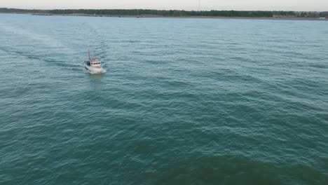 Aerial-view-of-a-coastal-fishing-boat-sailing-in-the-calm-Baltic-sea,-fisherman-dropping-fishing-net-in-the-water,-sunny-day,-distant-white-sand-beach,-wide-drone-shot-moving-backward