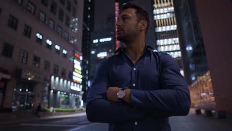 Ambitious-Young-European-Man-Standing-by-the-Road-at-Night-with-Crossed-Arms-Turning-Head-from-Left-to-Right-Enjoying-HIgh-rise-Towers-in-Seoul-Business-District