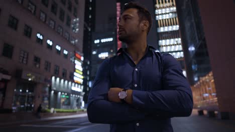 ambitious young european man standing by the road at night with crossed arms turning head from left to right enjoying high-rise towers in seoul business district