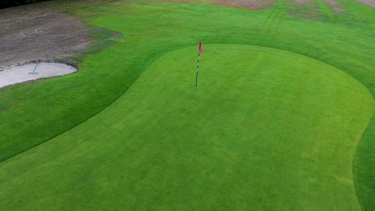 Aerial drone flies forward toward a golf flag on the green, then tilts downward into a top-down view above the pole and hole. Great stock clip for golf, sports, or environmental themes
