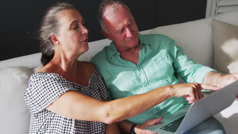 Caucasian-senior-couple-sitting-on-sofa-talking-in-the-sun-using-laptop