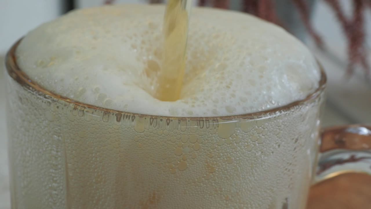 The tip of a crystal beer glass in focus as the beer is being poured, and the frothy foam fills the glass full in slow motion