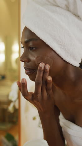 video vertical de una mujer afroamericana sonriente usando una toalla aplicando crema facial en el baño