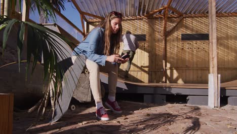 video of happy caucasian female skateboarder resting and using smartphone