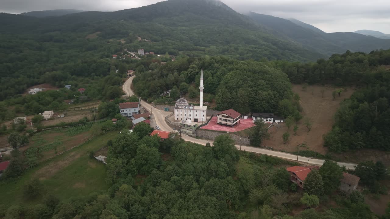Bulutlu bir günde yeşil tepelerle çevrili Mecidiye köyündeki geleneksel bir Türk camisinin havadan dairesel çekimi.
