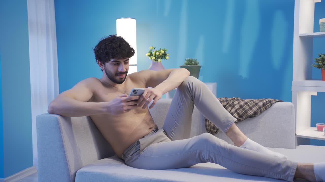 un joven guapo con un cuerpo en forma y en forma está acostado felizmente en el sofá, mirando su teléfono.