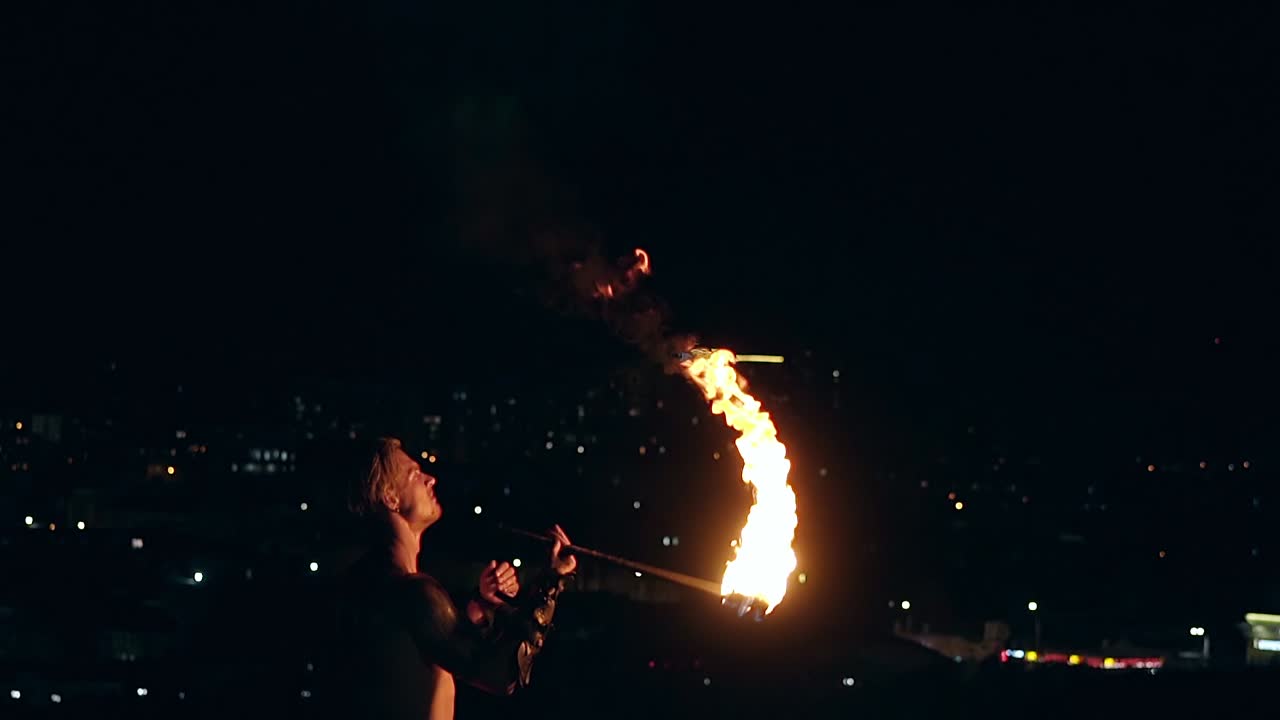 Young blond male breathes out large stream of fire with torch on black background Slow motion shot