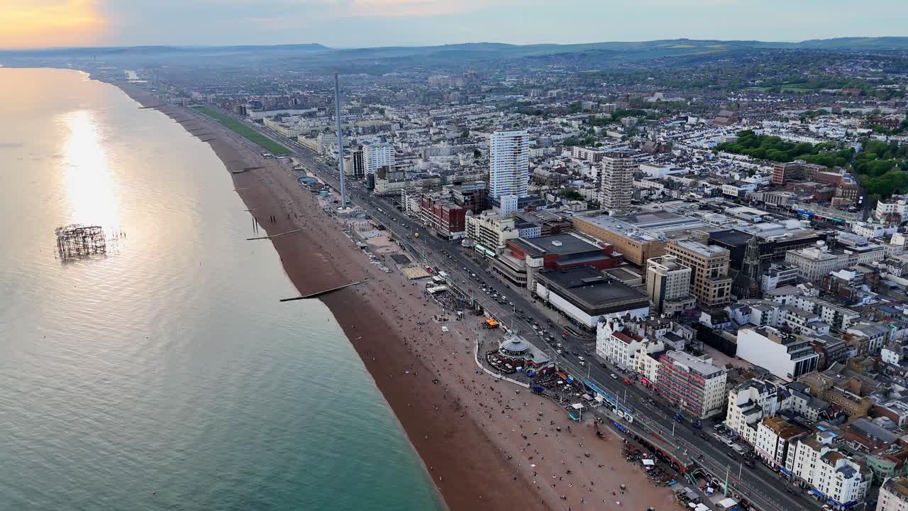 Stunning drone footage of Brighton seafront with busy beach, historic piers, and South Downs backdrop. Perfect for UK coastal, travel, tourism, and stock video content.