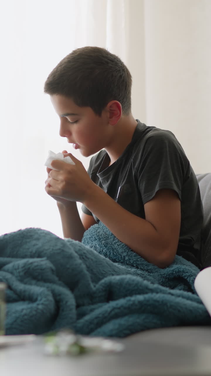 Bambino malato che starnutisce in un fazzoletto sul divano, coperto da una coperta, che mostra stanchezza vicino alla finestra con una tazza di vetro sul tavolo, che si sente male e ha bisogno di riposo per riprendersi dal raffreddore
