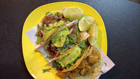 slow motion overhead shot of a yellow plastic plate with a assorted traditional mexican tacos served with slices of lime and guacamole on top