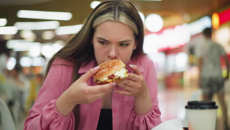 lady in pink dress smells burger with displeasure, showing disgust after dropping it, she reaches for a napkin to clean her hand while people in the background continue eating in a blurred view