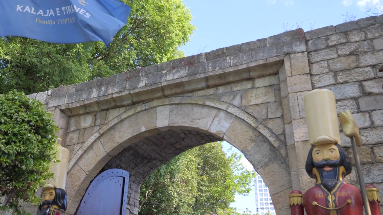 Laag standpunt van de stenen boogpoort van Kalaja e Tiranës, geflankeerd door houten Ottomaanse soldatenfiguren en een blauwe vlag in Tirana