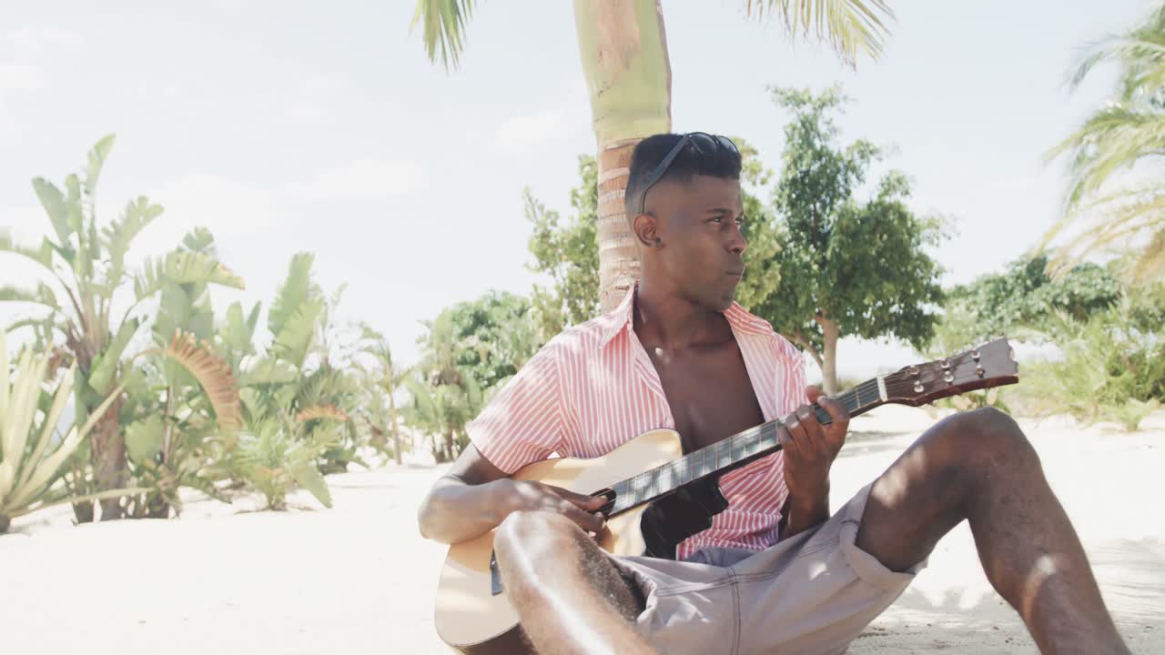 feliz hombre afroamericano sentado debajo de un árbol, tocando la guitarra en una playa soleada, cámara lenta
