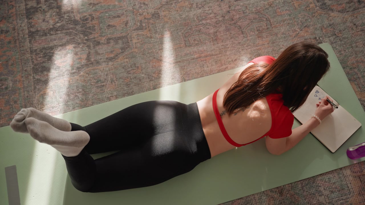 Donna sul tappetino che scrive un diario, vista dall'alto di un defaticamento post-allenamento a casa su un tappetino da yoga, quaderno accanto a borraccia e telefono, luce solare che filtra dalla finestra sul tappeto, recupero rilassato e fitness.