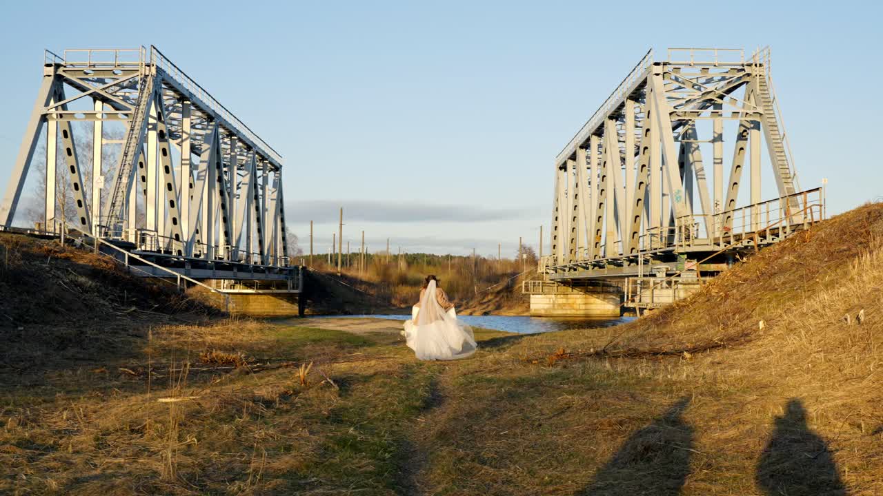 A bride in a white dress runs away from the camera, with two bridges and sunset light in view.