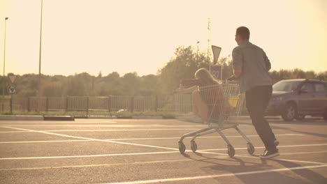 back view. cheerful young couple in love man and woman laughing and having fun while riding carts in supermarket parking in slow motion at sunset.