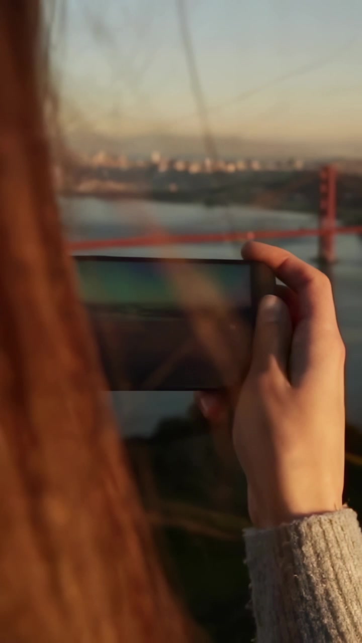 Vertical video: Sun urging woman holding phone at bay hair falling touching sleeve framing bridge