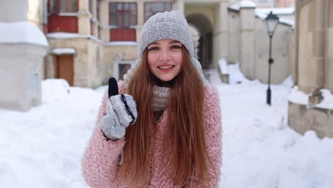 cheerful positive young woman smiling to camera, showing thumb up while standing outdoor in winter