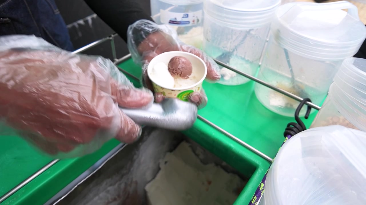 A vendor wearing plastic gloves scoops chocolate and vanilla ice cream into a paper cup.