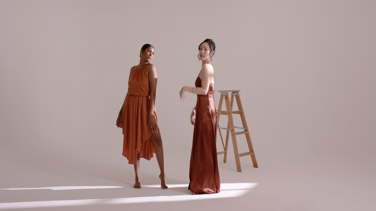 Indian and Chinese female models touching shoulder, shifting poses in studio with ladder for shoot