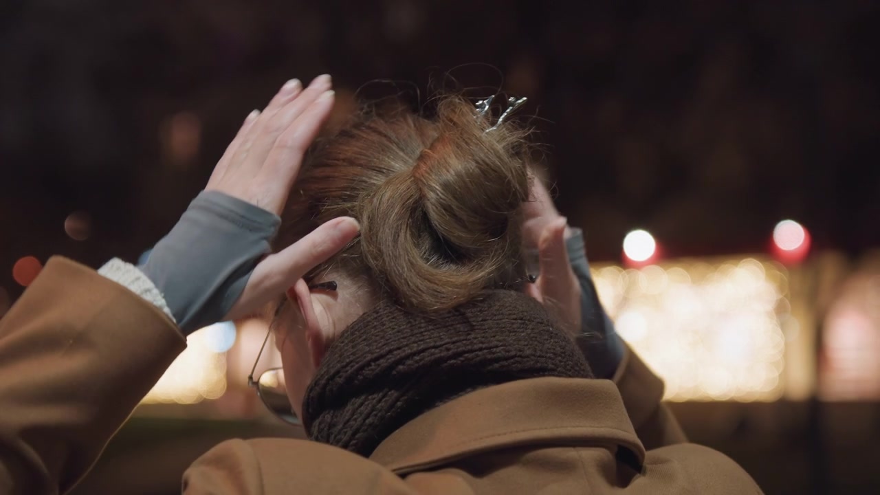 Evening park portrait of white woman adjusting wool scarf and hair bun, city bokeh lights, cold breath, thoughtful mood, glasses, brown coat, hands smoothing hair, candid rear profile
