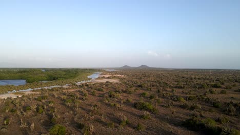 un avión no tripulado se mueve sobre una vasta extensión de tierra revelando una gran masa de agua en el lado izquierdo, dos montañas en el horizonte, como esta hermosa pradera en porlamar, isla margarita, venezuela