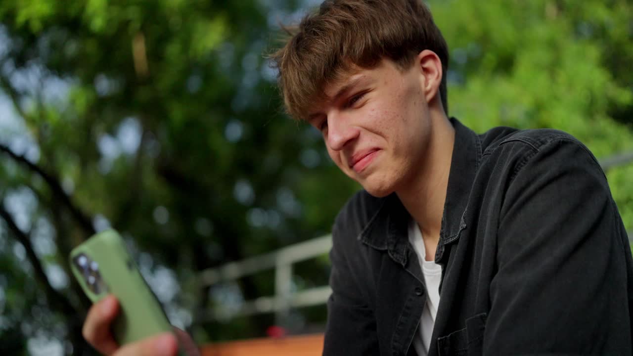 젊은 남자가 공원에서 앉아서 드폰을 쳐다보며 미소 짓고 있다.