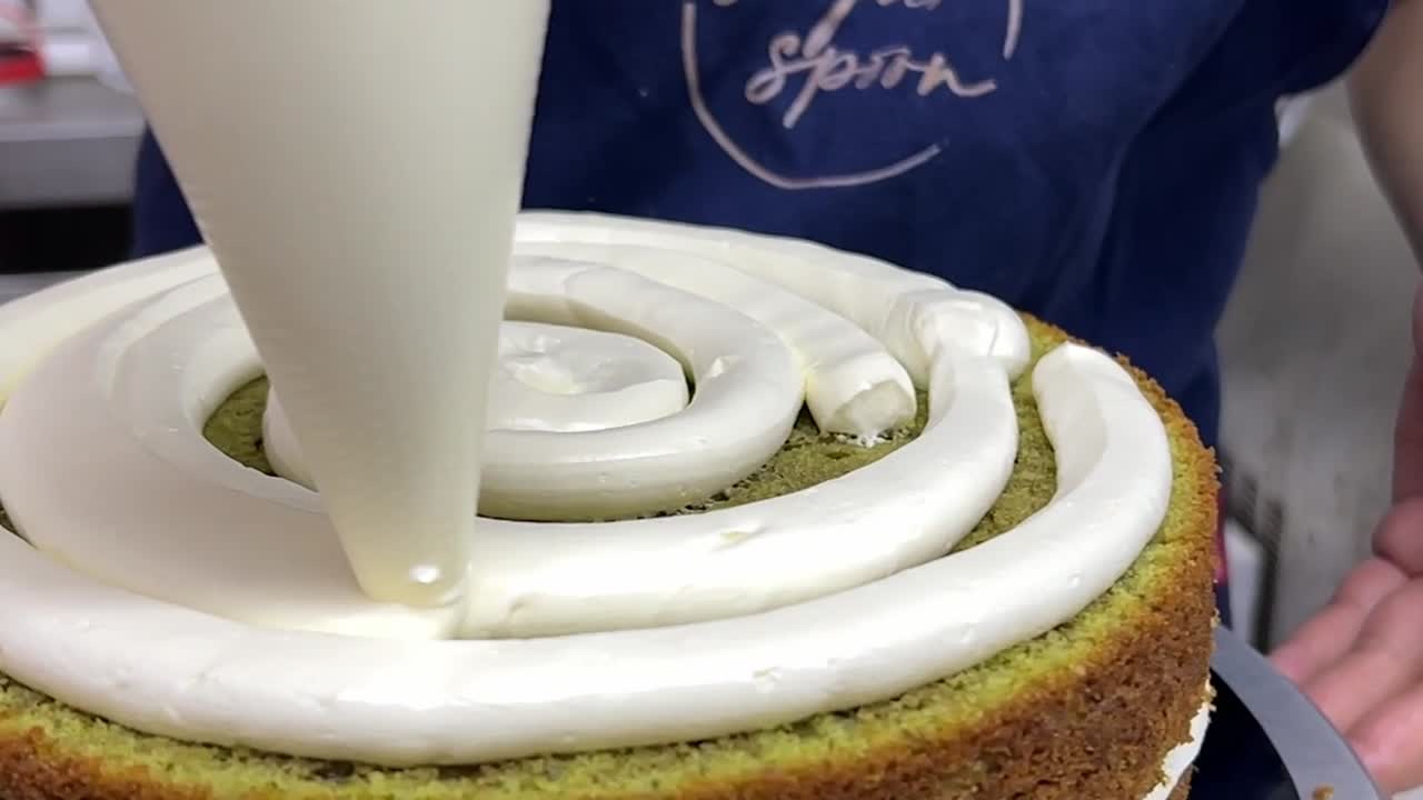 Close up shot of pastry chef using pipping bag to frost buttercream meringue in the middle layer of matcha green tea flavor fluffy sponge cake and gently spin the turntable