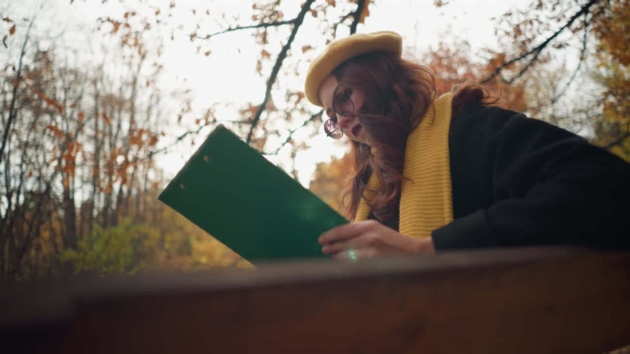 dama elegante sentada centrada en su libro con una portada verde, rodeada de hojas secas de otoño con la luz del sol brillando a través, en un entorno de parque sereno