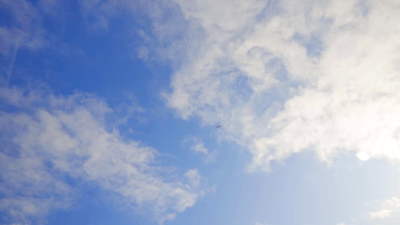 Extreme low angle looking at plane flying through blue and cloudy sky in summer. Rotating to keep it in frame. Slight lens flare.