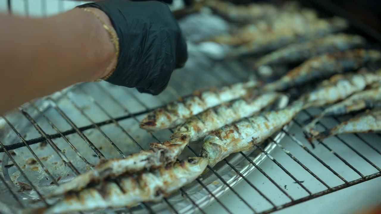 Persona con i guanti che griglia piccoli pesci su un barbecue, preparando sardine pronte da mangiare per street food o una cena estiva.