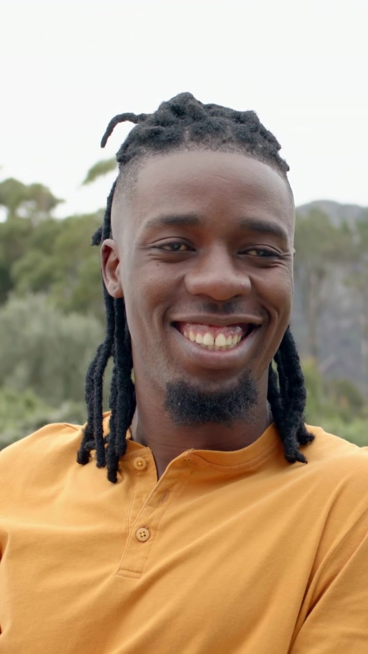 Video verticale: uomo sorridente che si gira verso un altro che entra nell'inquadratura, parla in un parco, maglietta henley arancione