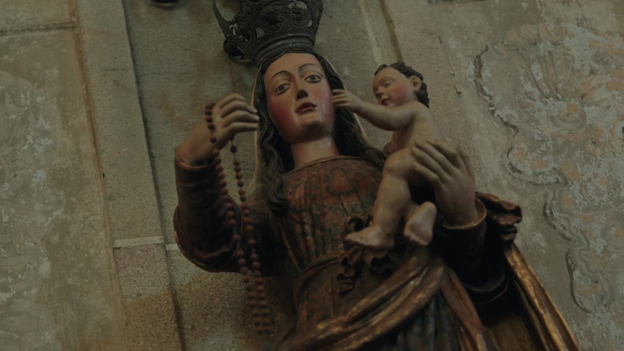 Estátua esculpida da Virgem Maria coroada segurando o Menino Jesus e um rosário dentro de uma igreja