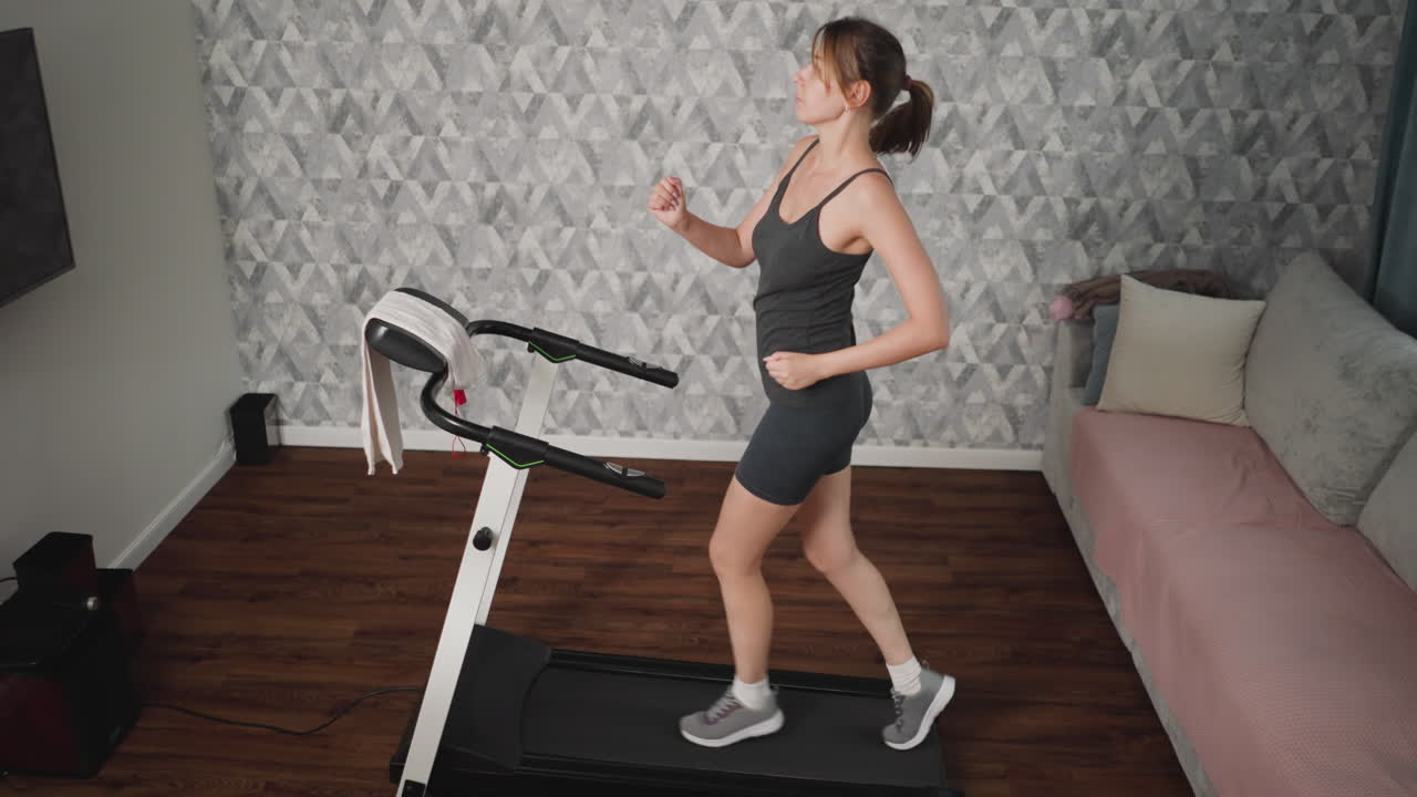 Determined mum running indoors on treadmill with focused expression, swinging arms and ponytail moving mid-stride, dressed in dark sportswear, towel draped on handlebar