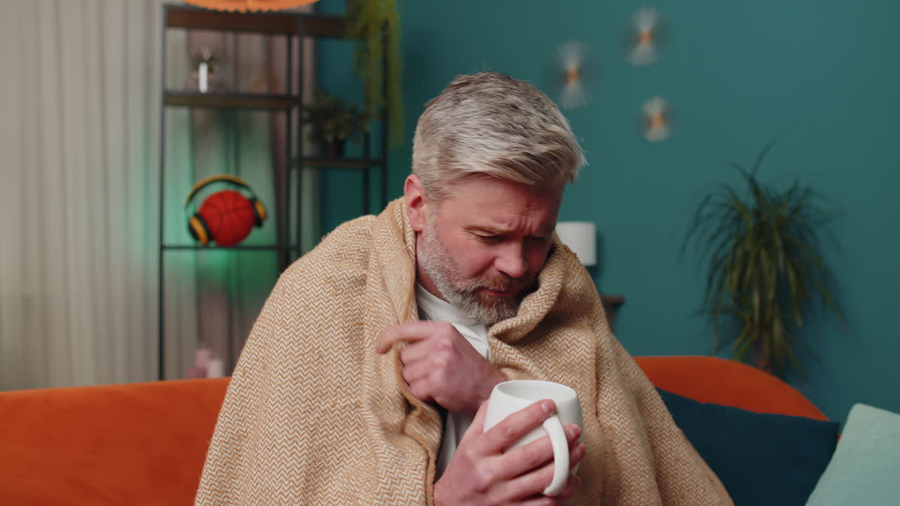 Sick unhealthy man wrapped in plaid sit alone shivering from cold on sofa drinking hot tea at home