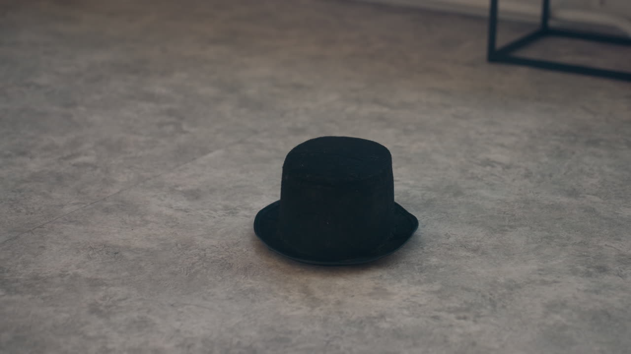 black hat tossed onto textured gray concrete floor near iron chair under diffused window light with dust particles swirling, subtle shadow contrast and minimal studio backdrop