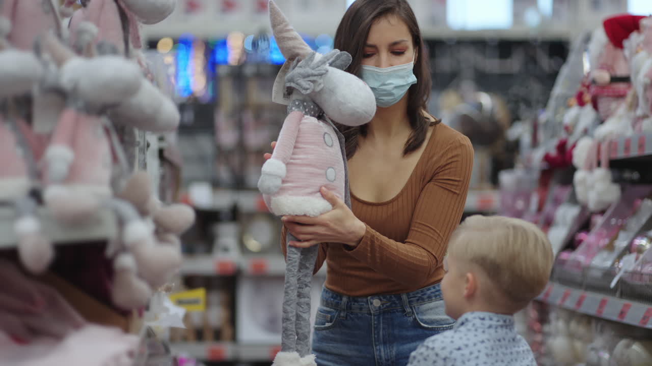 una madre con una máscara protectora toma un juguete del estante de la tienda y se lo muestra al niño