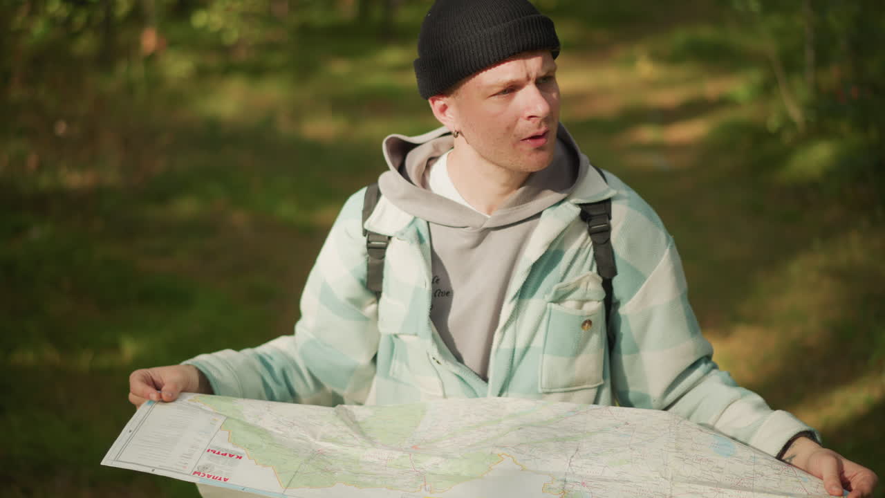 white hiker studying paper map outdoors, focused route planning among sunlit trees, backpack on shoulders, beanie hat, folded map held wide, soft foliage background, peaceful exploration mood