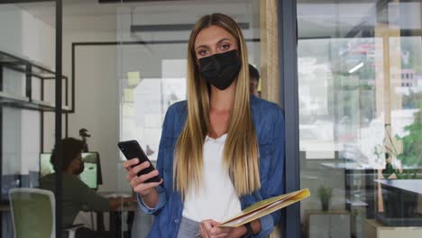 Portrait-of-caucasian-businesswoman-wearing-face-mask-using-smartphone-in-office