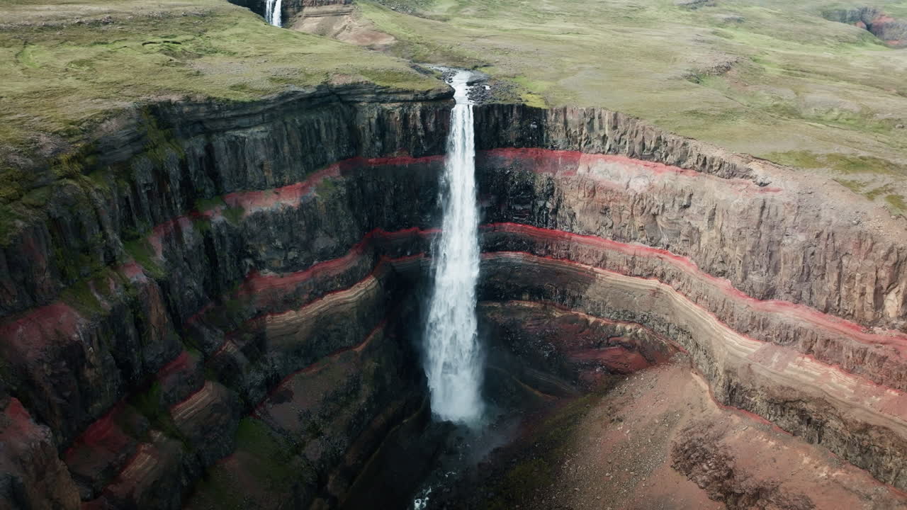 Wide aerial drone shot of Hengifoss Waterfall in motion, Iceland, sunny day, green grass, red clay. Part 1.