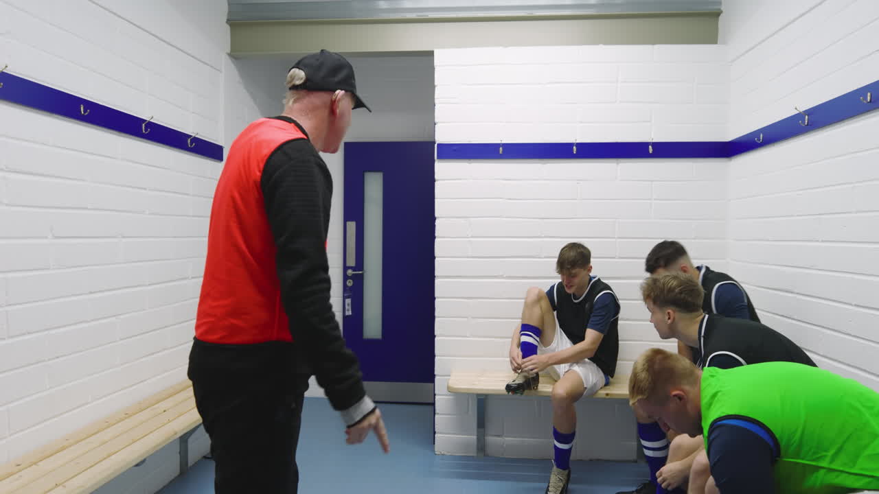 Male soccer players wearing black vests and tying shoes sitting on bench in changing room with coach